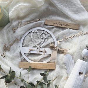 May include: A white wooden baptismal decoration with a heart and cross design, and the words "zur Taufe". Additional wooden cutouts say "zur Kommunion", "Firmung", and "zur Konfirmation". A beige ribbon is tied to the decoration.