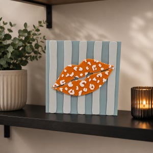 May include: A square decorative sign featuring a light blue background with vertical white stripes. Centered on the sign is a three-dimensional orange lip shape with a white leopard print pattern across the entire surface of the lips.
