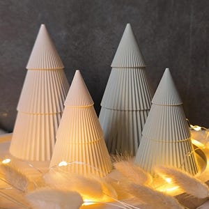 Peut inclure: Quatre arbres de Noël décoratifs en forme de cône, dans des tons de blanc et de gris. Chaque arbre présente un design côtelé et est orné d'une fine bande dorée. Les arbres sont disposés avec des guirlandes lumineuses et des éléments décoratifs blancs et duveteux.