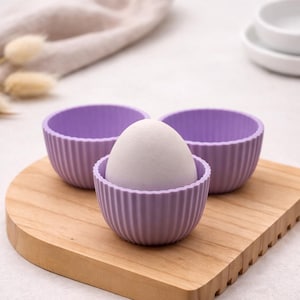 Może przedstawiać: Trzy lawendowe silikonowe foremki do pieczenia z żebrowanym wzorem, z których jedna zawiera biały przedmiot w kształcie jaja, na drewnianej desce do krojenia. Foremki są zgrupowane, sugerując motyw pieczenia lub kuchni.