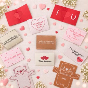 May include: A collection of rectangular greeting cards and paper cutouts featuring various heart shapes, otter illustrations, and German text phrases including I U, Jemand liebt dich, and Für immer, arranged on a light surface with small white flowers.