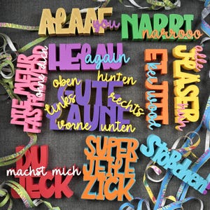 May include: A collection of colorful, die-cut words and phrases, including "ALAAF," "NARRI," and "SUPER ZICK," in various fonts and colors. The words are scattered on a gray surface with decorative ribbons.