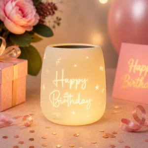 Puede incluir: Un portavelas de cumpleaños blanco roto y brillante con la inscripción "Happy Birthday" en letra cursiva. El portavelas está decorado con pequeñas estrellas y líneas onduladas. Una caja de regalo y flores rosas están en el fondo.