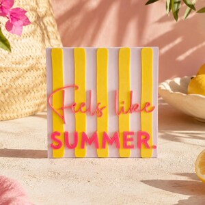 Puede incluir: Un letrero cuadrado con rayas verticales amarillas sobre un fondo morado claro. El texto "Feels like SUMMER." aparece en rosa, con "Feels like" en letra cursiva y "SUMMER." en letras mayúsculas negritas y sin remates.