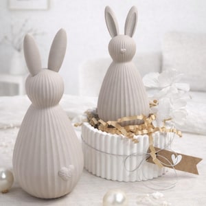 Könnte beinhalten: Zwei beige Keramik-Hasenfiguren. Ein Hase steht allein, der andere sitzt auf einer weißen, herzförmigen Schachtel mit zerknittertem Papier. Die Hasen haben lange Ohren und ein geripptes Körperdesign. Ein kleines Herzdetail befindet sich am Hinterteil des Hasens.