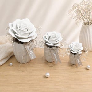 May include: Three white rose floral arrangements in textured grey pots. Each arrangement includes baby's breath and a decorative tag. The largest pot has a tag that reads "Best Mama". The other two have tags that read "Für dich".