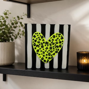 May include: A square decorative sign featuring a vertical black and white striped pattern. Centered on the sign is a large, bright neon yellow heart shape with a black leopard print pattern covering its entire surface.