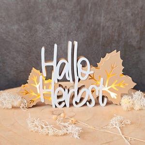 Könnte beinhalten: Ein Holzschild mit den Worten "Hallo Herbst" in weißer Schrift. Das Schild ist mit hellbraunen Blattausschnitten verziert und steht auf einer Holzoberfläche. Dekorative weißer Moos und getrocknete Stiele befinden sich im Vordergrund.