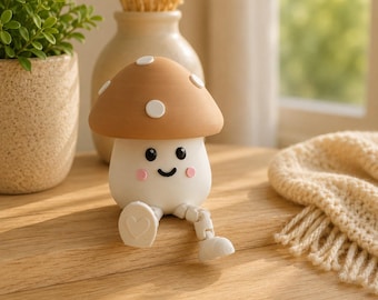 Puede incluir: Una pequeña figura de hongo con un sombrero color canela con puntos blancos, un tallo blanco y una cara sonriente con mejillas rosadas. La figura tiene piernas colgantes con pies blancos, uno de los cuales muestra un corazón en la suela.