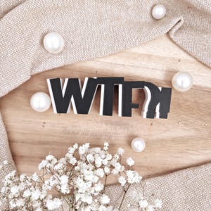 Puede incluir: Letras de madera en blanco y negro que deletrean "WTF" sobre una superficie de madera, rodeadas de perlas blancas y una tela beige. Flores blancas en la esquina inferior izquierda.