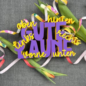 May include: A purple and yellow cutout with the German phrase "Gute Laune" (Good Mood) is surrounded by colorful ribbons and tulips. The words "oben, links, vorne, unten, hinten, rechts" are also present.