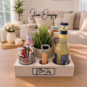 Può includere: Un vassoio di legno bianco con scomparti, contenente una bottiglia di soda, snack, una pianta e un apribottiglie. Il vassoio presenta le scritte "Zum Einzug" e "Bott & Salz".