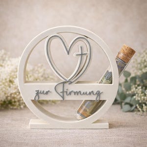 May include: A white, heart-shaped decorative piece with a cross, a circular frame, and the text "zur Firmung". A small glass vial with a cork stopper holds rolled-up currency. The design is simple and elegant, suitable for a religious gift.