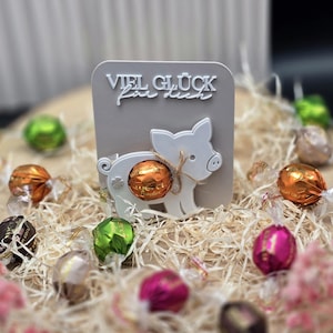 Pode incluir: Um cartão cinza com o texto "VIEL GLÜCK für dich" e um recorte de porco branco segurando um chocolate embrulhado. Vários chocolates embrulhados em laranja, verde, rosa e marrom estão espalhados ao redor do cartão em uma cama de palha.