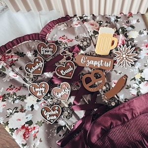May include: A collection of German-themed keychains and accessories, including heart-shaped charms with German phrases, a pretzel, a beer mug, and a flower. The items are arranged on a floral patterned fabric with burgundy accents.