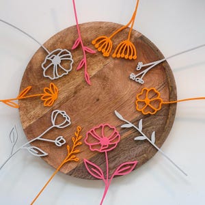 Puede incluir: Una colección de recortes de flores coloridas y estilizadas en tonos rosa, naranja y gris, dispuestas sobre una tabla redonda de madera. Las flores tienen tallos delicados y diseños intrincados de pétalos, creando una exhibición decorativa y alegre.