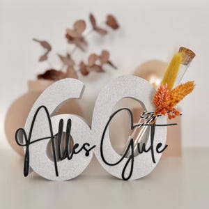 Könnte beinhalten: Eine dekorative '60' mit den Worten 'Alles Gute' in schwarzer Schrift. Die Zahl ist hellgrau mit strukturierter Oberfläche. Ein kleines Glasröhrchen mit getrockneten orangefarbenen und gelben Blumen ist befestigt.