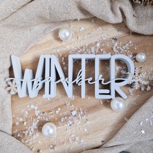 Pode incluir: Uma placa de madeira azul clara que diz "Winter" com "December" em itálico. A placa é decorada com flocos de neve, pérolas e neve falsa em uma superfície de madeira. Um tecido de cor neutra é drapeado no fundo.