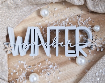 Puede incluir: Un letrero de madera azul claro que dice "Winter" con "December" en cursiva. El letrero está decorado con copos de nieve, perlas y nieve artificial sobre una superficie de madera. Una tela de color neutro está drapeada en el fondo.