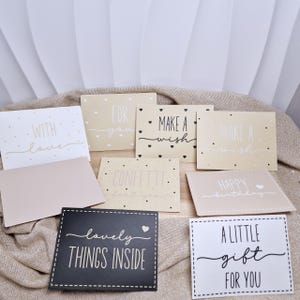 Puede incluir: Una colección de tarjetas de felicitación con varios mensajes en fuente cursiva. Las tarjetas son de colores neutros, con frases como "Con amor", "Para ti" y "Pide un deseo". Algunas tarjetas tienen pequeños acentos de corazón y puntos.