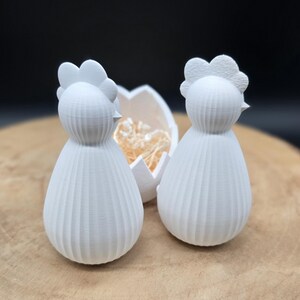 May include: Two white chicken figurines with textured bodies and detailed crests, on a wooden surface. Behind them is a cracked eggshell filled with straw. The figurines are likely for Easter or spring decor.