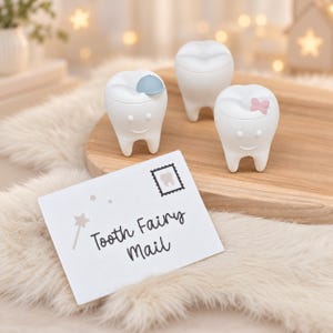 Könnte beinhalten: Drei weiße, zahnförmige Behälter mit lächelnden Gesichtern, einer mit einer blauen Mütze und einer mit einer rosa Schleife. Eine weiße rechteckige Karte zeigt den Text Tooth Fairy Mail, eine kleine Sternstabgrafik und eine Briefmarke.