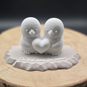 May include: A light gray penguin figurine holding a heart. The figurine has the text "Du bist mein für immer" on the base. The penguins are on a gray base with a textured, geometric design.