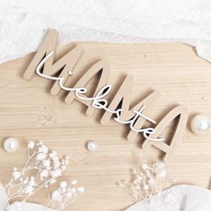 May include: A wooden sign with the word "MAMA" in a beige color, with the word "liebste" in white script overlaying it. The sign is on a light wood surface, with white flowers and pearls.