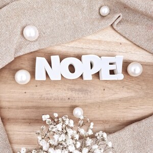 Puede incluir: Una superficie de madera con la palabra "NOPE!" en letras blancas. La palabra está rodeada de perlas blancas, una tela beige y un pequeño racimo de flores blancas.