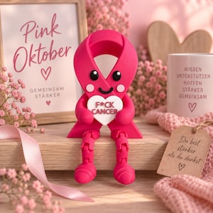 Op de afbeelding: Een felroze lintfiguur met een glimlachend gezicht, zwarte ogen en roze wangen, die een wit hartvormig bordje vasthoudt met de tekst F*CK CANCER. Het figuurtje heeft beweegbare benen en voeten en staat in een omgeving met roze bloemen.