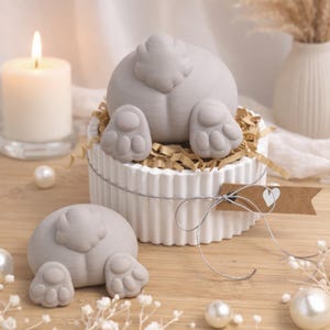Könnte beinhalten: Zwei graue Hasenpopo-Figuren mit Pfoten-Details, eine davon in einem weißen, gerippten Behälter mit geschreddertem Papier und einem kleinen Etikett. Eine brennende Kerze, Perlen und dekorative Blumen im Hintergrund. Die Figuren stehen auf einer Holzoberfläche.