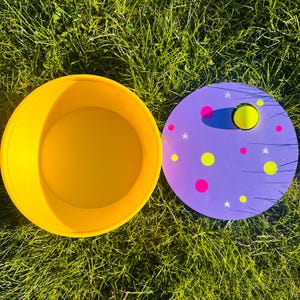 Puede incluir: Un recipiente de plástico amarillo junto a una tapa morada decorada con círculos rosas y amarillos, estrellas y una esfera amarilla. Los artículos están sobre hierba verde.