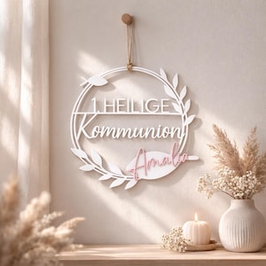 Op de afbeelding: Een witte houten krans met de woorden "1 HEILIGE Kommunion" en de naam "Amalia" in roze. De krans is versierd met bladeren en hangt aan een houten peg.