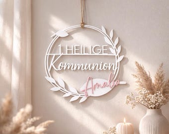 May include: A white wooden wreath with the words "1 HEILIGE Kommunion" and the name "Amalia" in pink. The wreath is decorated with leaves and hangs from a wooden peg.