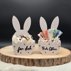 Könnte beinhalten: Zwei Hasen-förmige Osterkörbe aus hellem Holz, die jeweils ein anderes Geschenk enthalten. Einer enthält eine Kerze und der andere gefaltetes Geld. Die Körbe sind mit schwarzen Herzen und Text verziert.
