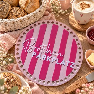Könnte beinhalten: Ein rundes, hellrosa Holzbrett mit vertikalen dunkelrosa Streifen und einer Lochreihe am Rand. Der Text "Brötchen PARKPLATZ" ist in Weiß aufgebracht, wobei "Brötchen" in Schreibschrift und "PARKPLATZ" in Blockbuchstaben dargestellt ist.