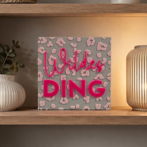 May include: A square decorative sign featuring a gray background with a raised, light pink leopard print pattern. The sign displays the word Wildes in a bright pink cursive script font above the word DING in bold, bright pink block letters.