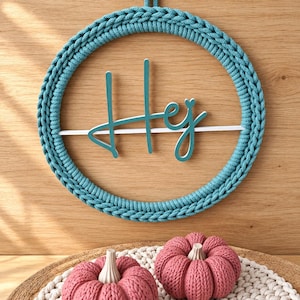 Könnte beinhalten: Ein kreisförmiger Wandbehang mit einem Rahmen aus blaugrünem Strickseil und einem mittigen weißen horizontalen Balken mit dem blaugrünen Schriftzug "Hej". Darunter befinden sich zwei kleine, runde, rosa Strickkürbisse.