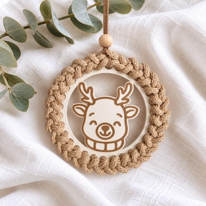 May include: A circular hanging ornament featuring a braided tan rope border surrounding a white wooden center. The center displays a brown line-art illustration of a reindeer face with antlers. A small wooden bead sits above the circle.