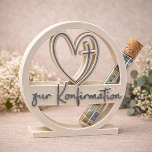 May include: A cream-colored Confirmation gift holder with a heart and cross design. The words "zur Konfirmation" are written across the front. Two glass vials with cork stoppers are inserted into the holder.