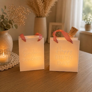 Könnte beinhalten: Zwei weiße Geschenktüten aus Papier mit rosa Bandhenkeln, die von innen beleuchtet sind. Eine Tüte zeigt den geprägten Text "Ein Jahr SCHÖNER" und die andere "Happy BIRTH DAY" in erhabener Schrift. Eine Glasflasche mit Korken steht darin.