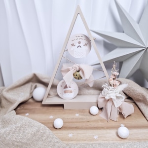 Peut inclure: Décoration de Noël en forme d'arbre en bois avec deux ornements ronds, une cloche et un arrangement floral. Les ornements comportent des illustrations et du texte. L'arbre est sur une surface en bois avec des boules décoratives blanches et une étoile.