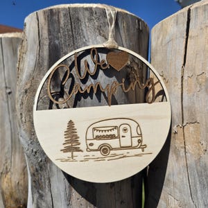 Könnte beinhalten: Runder Holzschild mit den Worten "We Love Camping" in einer Schriftart ausgeschnitten. Das Schild zeigt ein kleines Holzherz und ein eingraviertes Bild eines Wohnwagens und eines Baumes. Eine Schnurschlaufe zum Aufhängen ist angebracht.