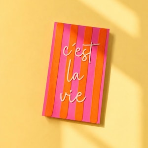 Puede incluir: Una tarjeta rectangular con rayas verticales en tonos alternos de rosa brillante y naranja. Centrado en la tarjeta se encuentra el texto en cursiva blanca c'est la vie, escrito en tres líneas. El diseño es plano y vertical.