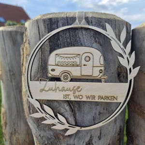 Könnte beinhalten: Holz-Wandbehang in Wohnwagenform mit dem Text "Zuhause ist, wo wir parken." Das Design umfasst einen kreisförmigen Rahmen mit Blättern und einem Herz an der Wohnwagentür. Ein klares Band ist zum Aufhängen befestigt.
