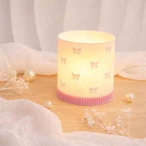May include: A cylindrical candle holder with a pink base and bow pattern. The candle holder is white with pink bows and a pink ribbed base. Pearls and white flowers are scattered around the candle holder.