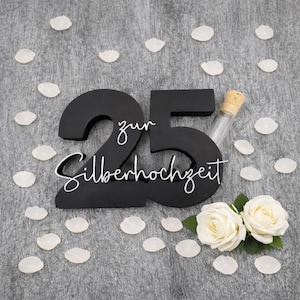 Könnte beinhalten: Eine schwarze Holznummer 25 mit den Worten "zur Silberhochzeit" in weißer Schrift. Das Bild enthält ein kleines Glasfläschchen mit Korken, weiße Rosenblätter und zwei weiße Rosen.
