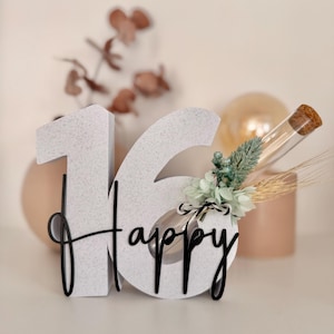 Könnte beinhalten: Ein dekoratives '16 Happy'-Schild mit gesprenkelter weißer Oberfläche. Das schwarze kursive 'Happy' ist an den Zahlen befestigt. Die '6' ist mit kleinen Blumen, Weizen und einem Glasfläschchen mit Korken verziert. Der Hintergrund enthält unscharfe dekorative Elemente.