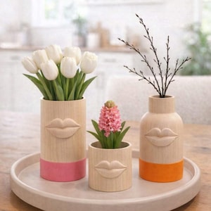 Könnte beinhalten: Drei Holzvasen mit geschnitzten Lippenmotiven, jede mit Blumen oder Zweigen. Die Vasen haben eine natürliche Holzoberfläche mit farbigen Böden in Rosa und Orange. Die Vasen sind auf einem runden Tablett angeordnet.