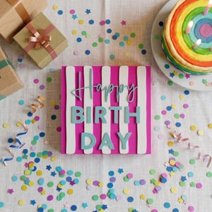Może przedstawiać: Kwadratowy znak w pionowe różowe i białe paski z napisem Happy BIRTH DAY jasnoniebieskimi literami 3D, otoczony rozrzuconym kolorowym konfetti, zapakowanymi pudełkami prezentowymi i tęczowym tortem z zapalonymi świeczkami.
